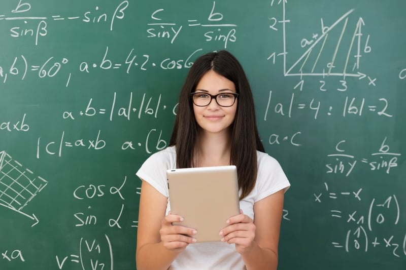 Clever attractive young female college student wearing glasses and holding a tablet computer in her hands standing in front of a chalkboard covered in equations in maths class smiling at the camera